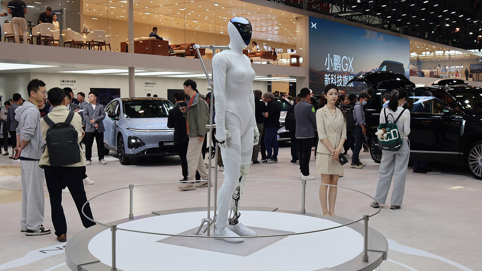 An Xpeng Iron humanoid robot stands on a display platform at the 2026 Beijing auto show, with Xpeng vehicles and visitors in the background.