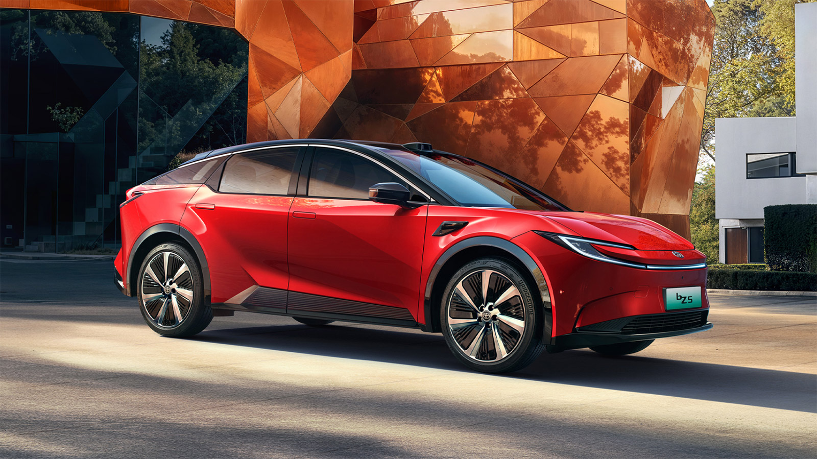 Red 2027 Toyota bZ5 electric coupe SUV parked outside a modern copper-clad building.