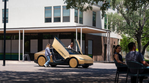 A gold-colored Tesla Cybercab parked in a plaza with its butterfly doors raised as passengers get into the vehicle.