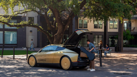 A gold-colored Tesla Cybercab parked under trees as a man loads a guitar case into its open rear cargo area.