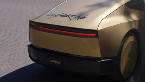Close-up of the rear of a gold-colored Tesla Cybercab, showing its full-width red light bar and Cybercab branding.