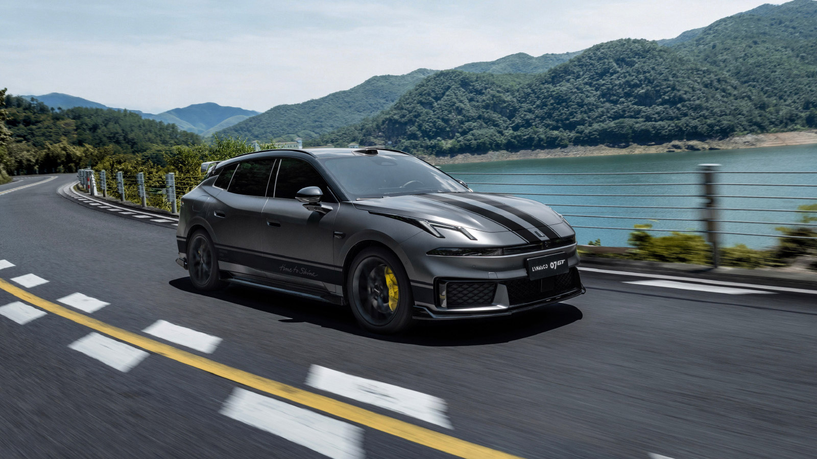 A gray Lynk & Co 07 GT with black racing stripes driving along a lakeside road, with forested mountains in the background.