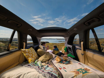Woman and child relaxing on bedding inside an Onvo L80, with the rear seats folded and the open tailgate overlooking a lake.