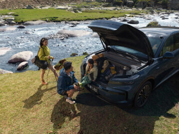 Blue Onvo L80 parked beside a rocky stream, with its front trunk open and a family gathered around hiking gear.