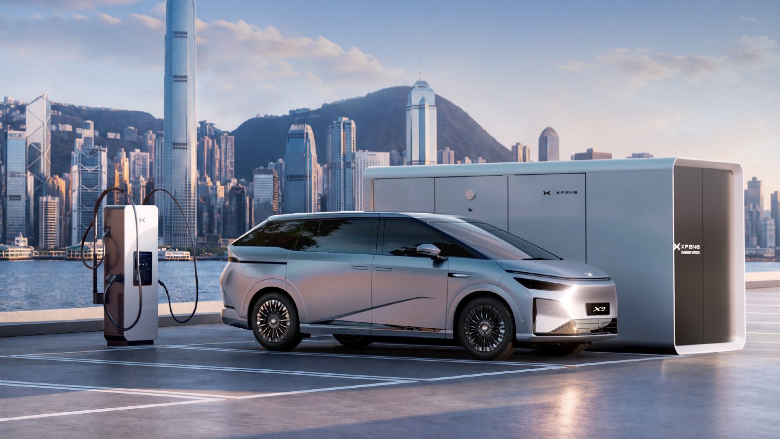 Silver Xpeng X9 MPV beside a charging unit and energy storage cabinet, with Hong Kong's skyline across the harbor.