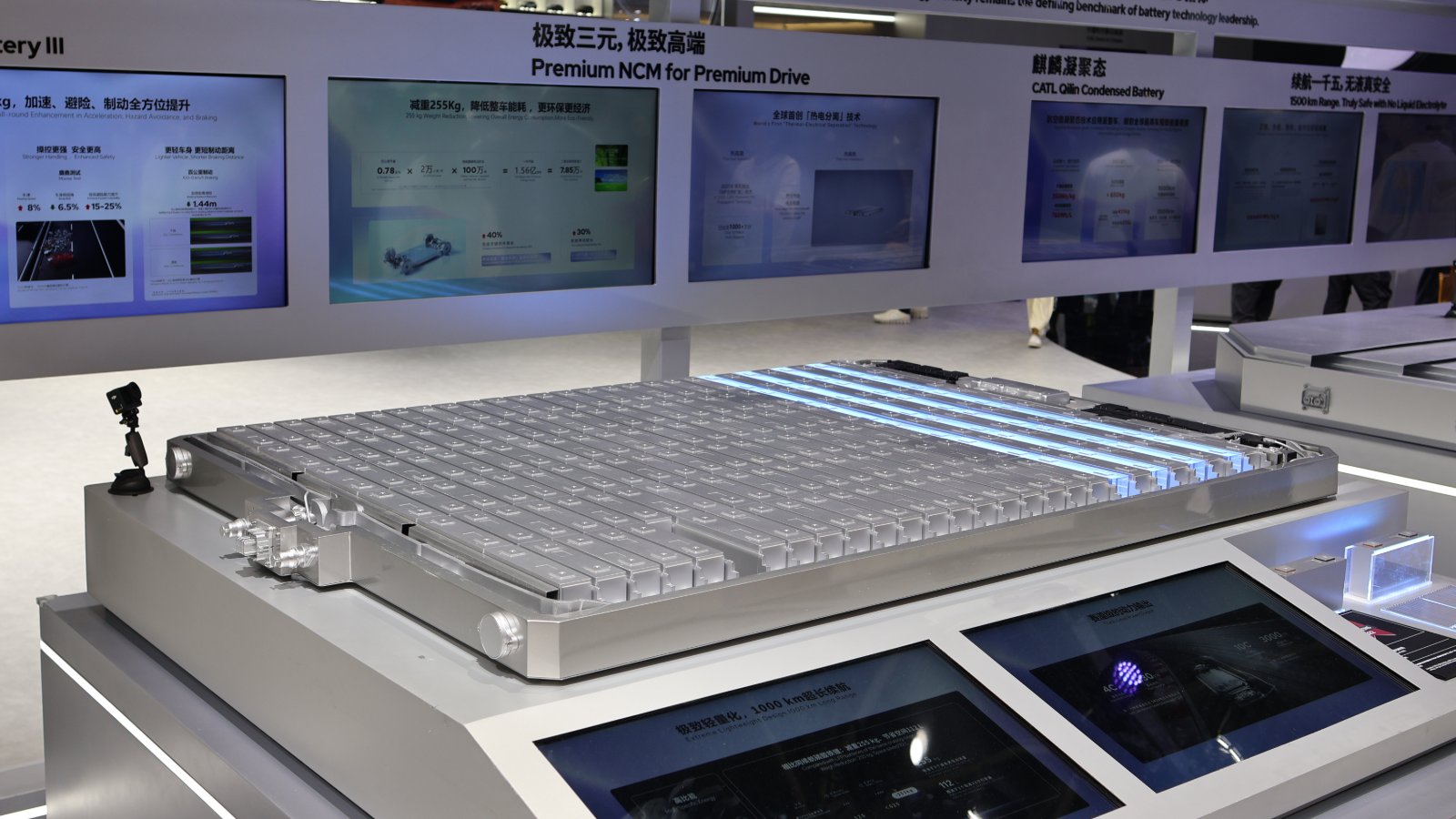 A CATL battery pack with rows of exposed prismatic cells and blue-lit cooling channels displayed on a stand at the Beijing auto show, with screens behind it showing CATL's "Premium NCM for Premium Drive" and Qilin condensed battery exhibits.