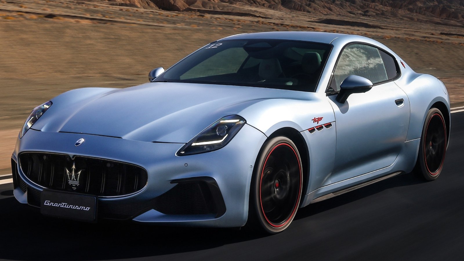 A light blue Maserati GranTurismo driving on a road, viewed from the front three-quarter angle.