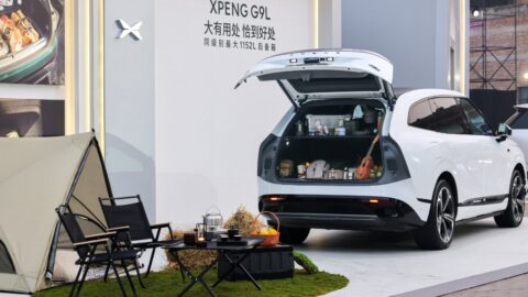 White Xpeng G9L with its tailgate open, showing luggage, camping supplies and a ukulele beside a tent and folding chairs.