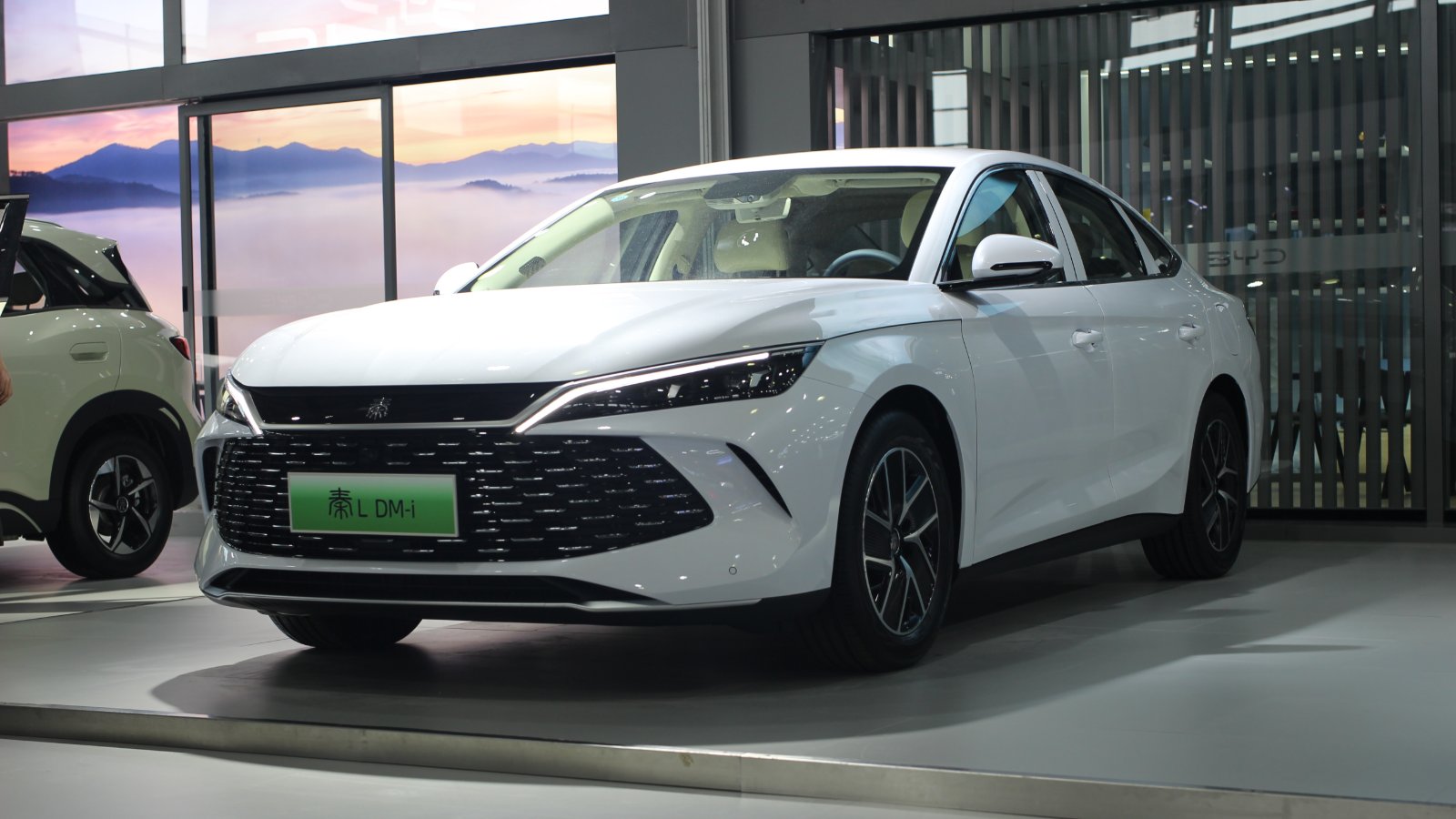 White BYD Qin L DM-i plug-in hybrid sedan on display indoors, viewed from the front and side.