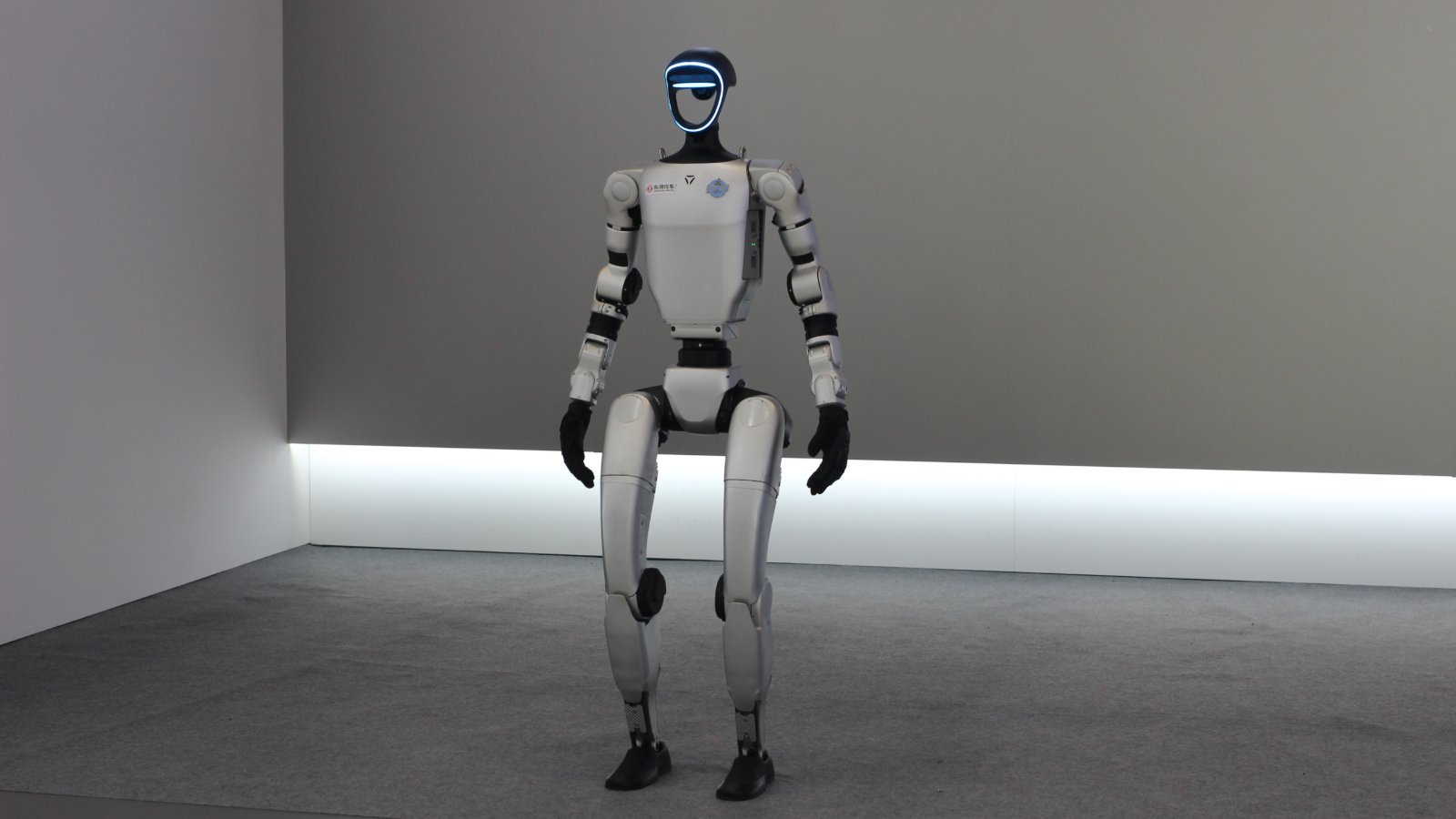 A white humanoid robot with black joints and a blue-lit visor stands upright on a grey carpeted floor in front of a dark grey wall at Dongfeng's exhibition booth, with the Dongfeng Motor logo on its chest.
