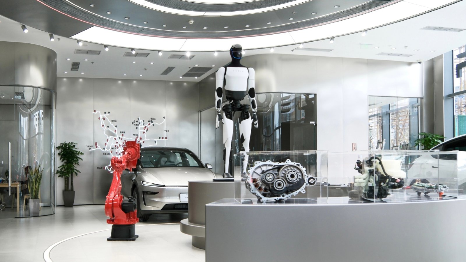 A Tesla Optimus humanoid robot stands on a display platform at a Tesla store in Beijing, next to a red industrial robot arm, a silver Model Y and drivetrain components in glass cases.