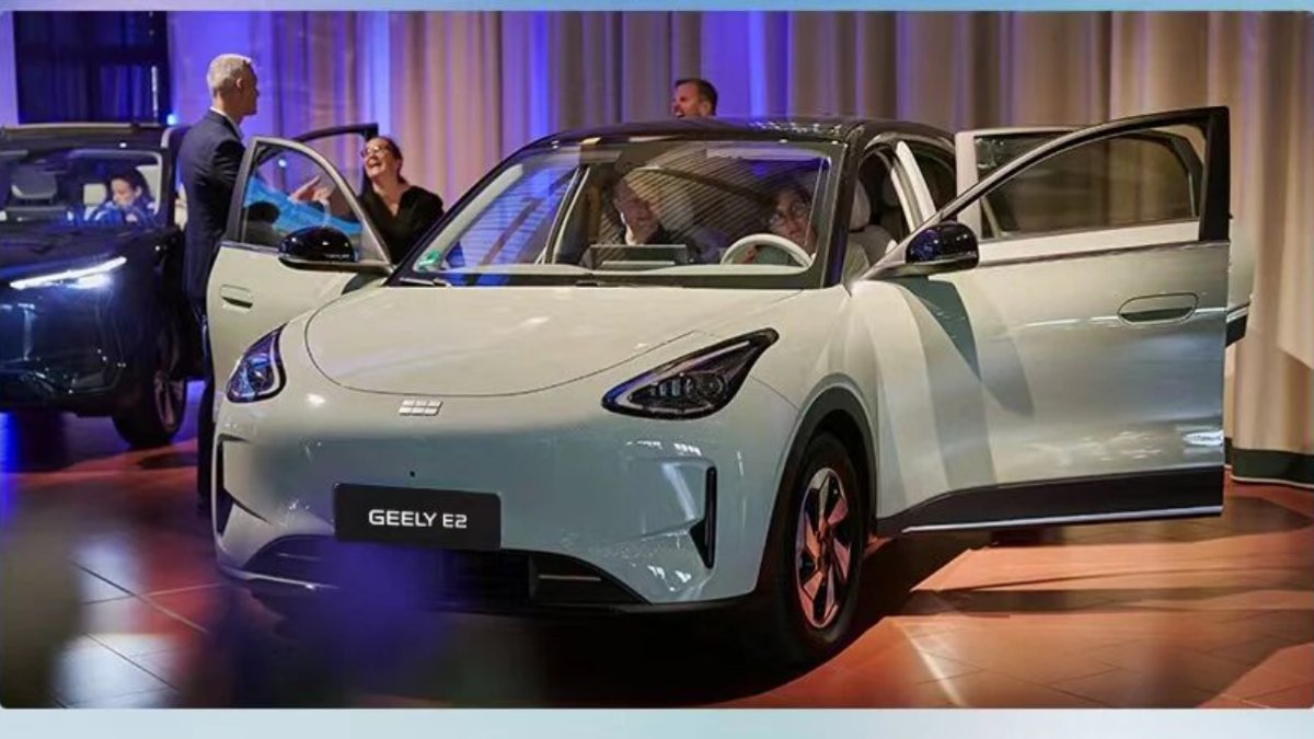 A pale green Geely E2 battery electric crossover displayed indoors with its doors open, as guests sit inside the car and examine the interior at Geely's launch event in Switzerland.