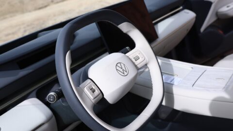 Close-up of the ID. UNYX 09's two-tone steering wheel with a dark blue rim, cream-colored center pad bearing the Volkswagen logo, and roller controls on the spokes, with a light-colored dashboard, wood-look trim and center console in the background.
