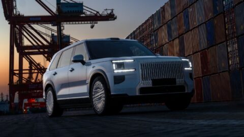 Front three-quarter view of a silver Zeekr 9X with illuminated headlights beside shipping containers and cranes.