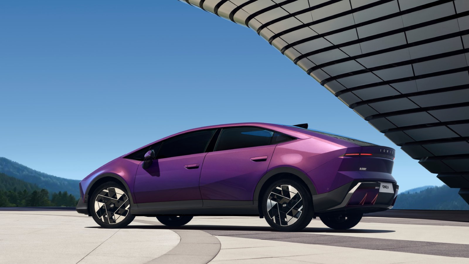 Rear three-quarter view of a purple Hyundai Ioniq V electric sedan parked under a modern architectural canopy, showing its sloping roofline and geometric-pattern wheels.