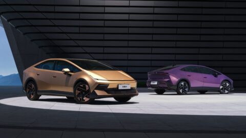 A gold Hyundai Ioniq V facing forward and a purple Ioniq V facing away, parked side by side in front of a dark slatted building facade.