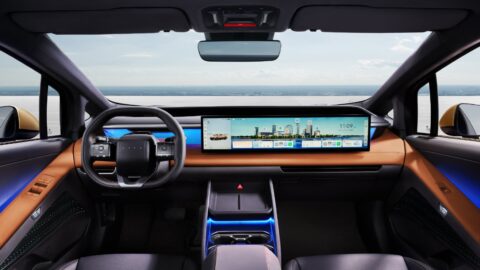 Front cabin of the Hyundai Ioniq V, showing a squared-off steering wheel, a 27-inch ultra-wide central touchscreen, orange dashboard trim, and blue ambient lighting.