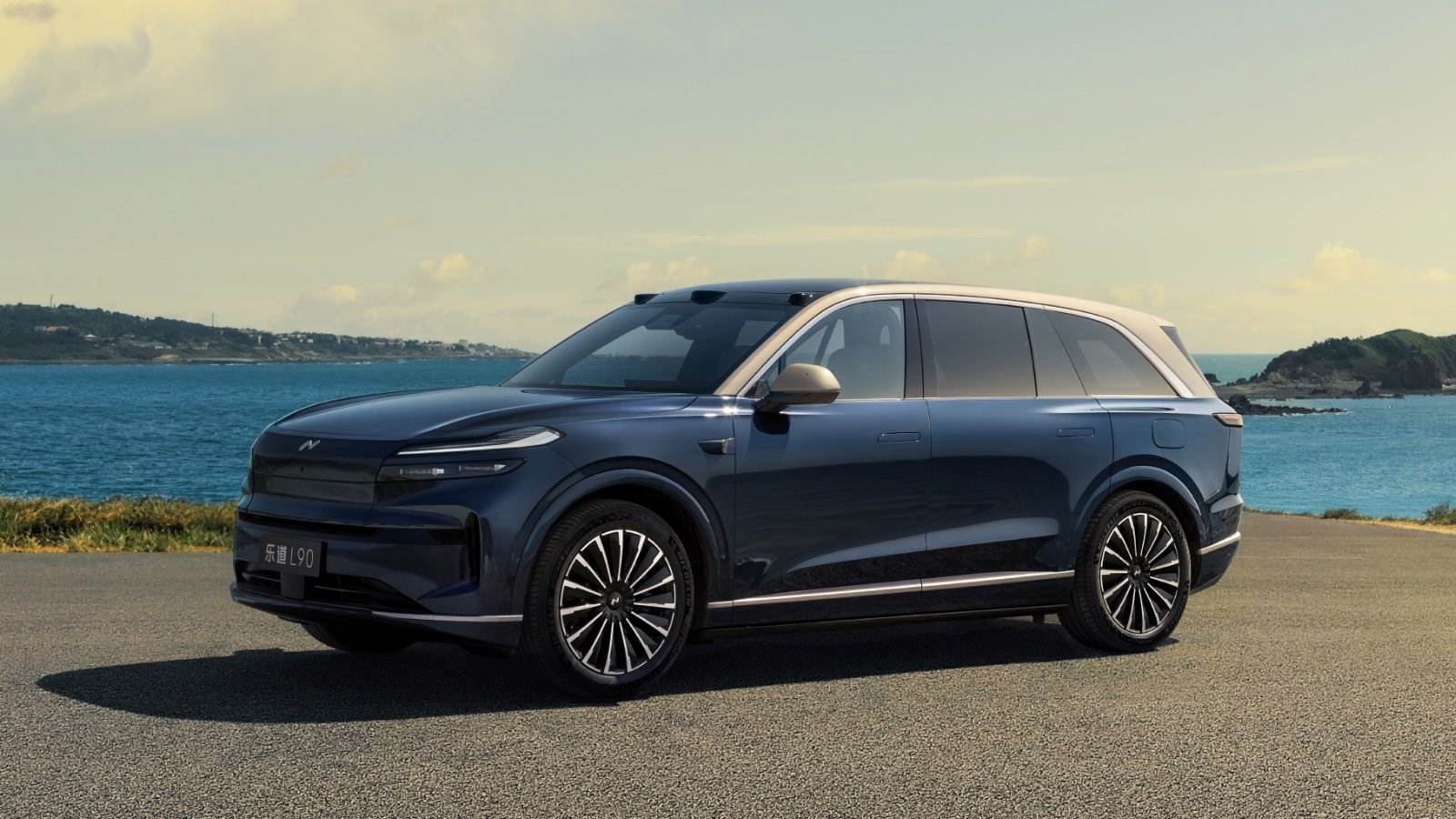 Front three-quarter view of the Onvo L90 with the Starry Gold design package, finished in dark Starry Blue with a gold-toned roof and titanium-colored trim, parked on a seaside road with the ocean and hills in the background.