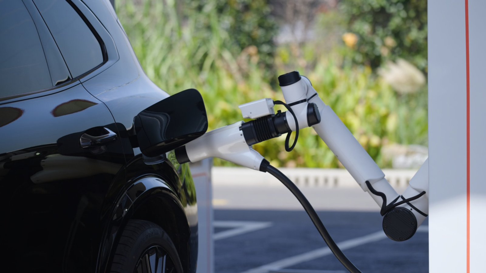 A white robotic charging arm plugs a connector into the charging port of a black electric vehicle parked outdoors, with green shrubs and trees in the background.