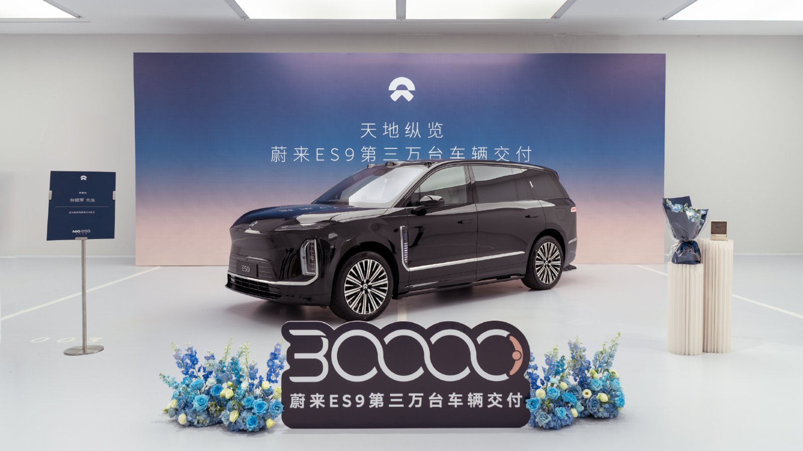 A dark-colored Nio ES9 displayed behind a 30,000-delivery milestone sign and blue floral arrangements.