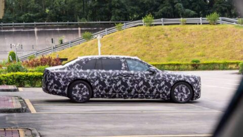 Side profile of a camouflaged Yangwang executive sedan prototype on a paved road beside a grassy slope.