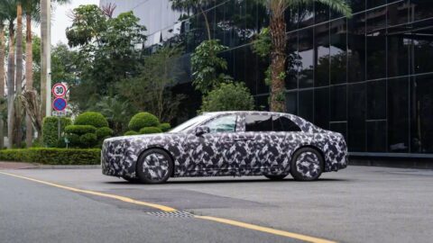 Camouflaged Yangwang executive sedan prototype outside a glass-fronted building, showing its long hood and large rear doors.