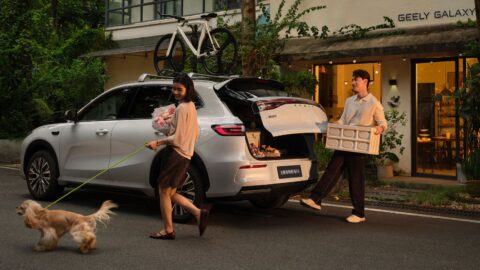 Geely Galaxy E5 with its rear liftgate open as a man carries a crate and a woman walks a dog nearby.