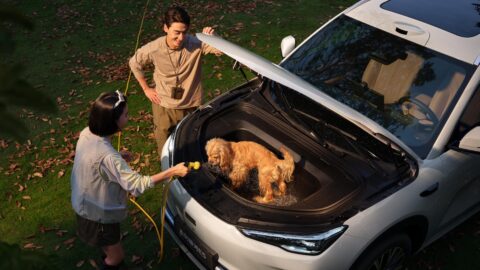 Dog sitting in the open front trunk of a Geely Galaxy E5 as 2 people stand beside the vehicle.
