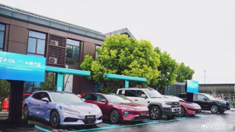 Sedans and SUVs parked beneath turquoise BYD flash chargers outside a highway service-area building.
