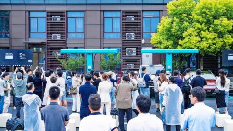 A crowd gathers around vehicles and turquoise BYD flash chargers at a highway service area during the station completion event.