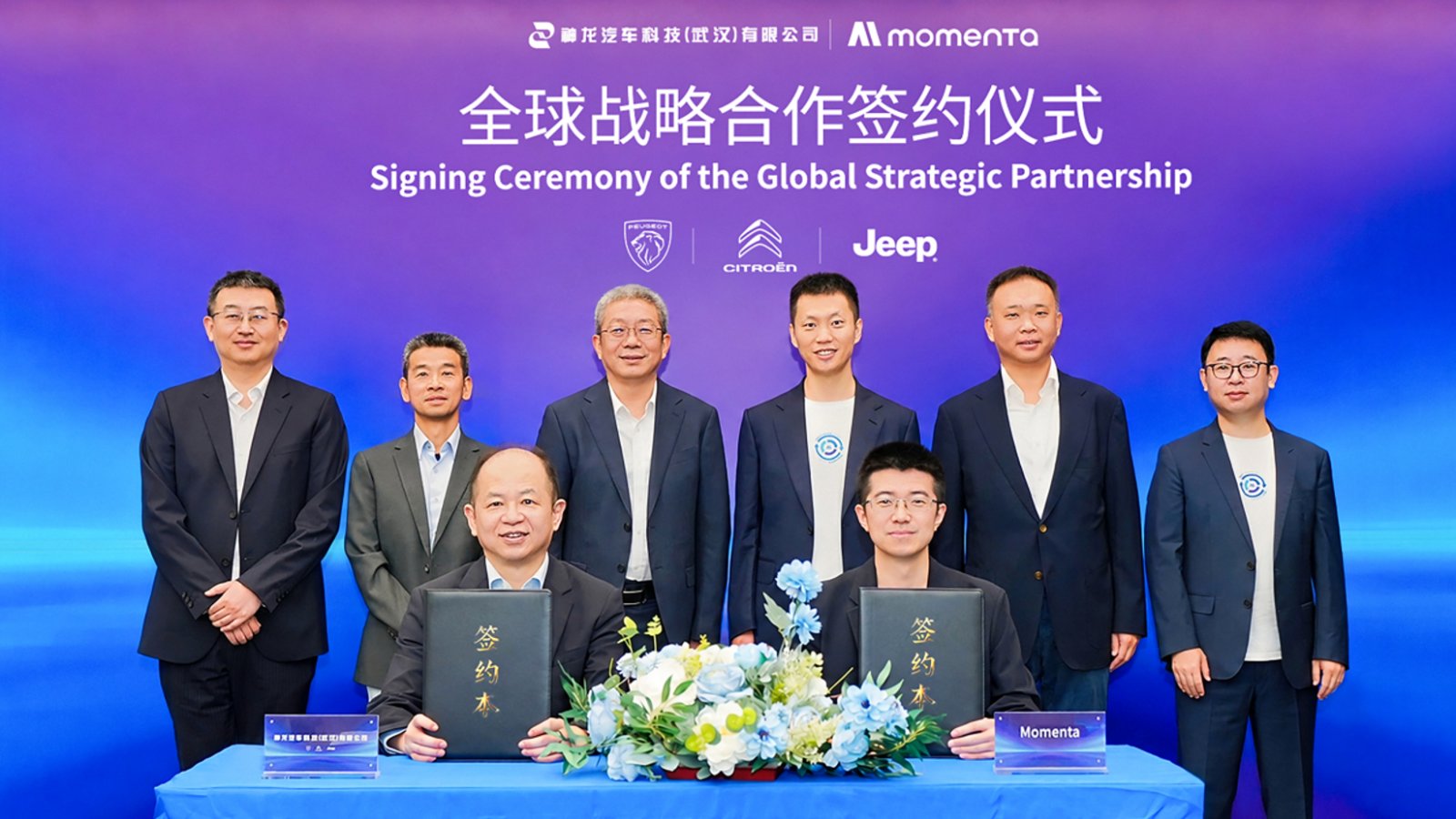 Momenta and DSAT representatives hold agreement folders at a global strategic partnership signing ceremony, with executives standing behind them.