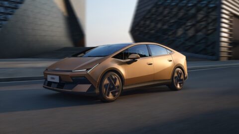 Gold Hyundai Ioniq V electric sedan driving past modern buildings, viewed from the front three-quarter angle.