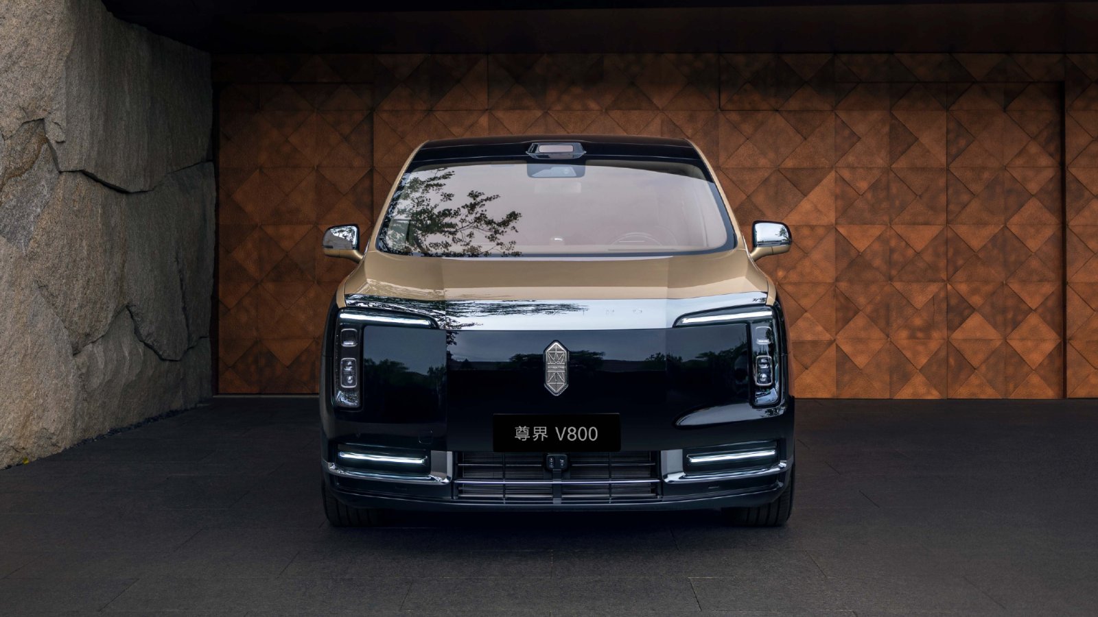 Front view of a gold-and-black Maextro V800 luxury MPV parked in front of a patterned wooden wall.
