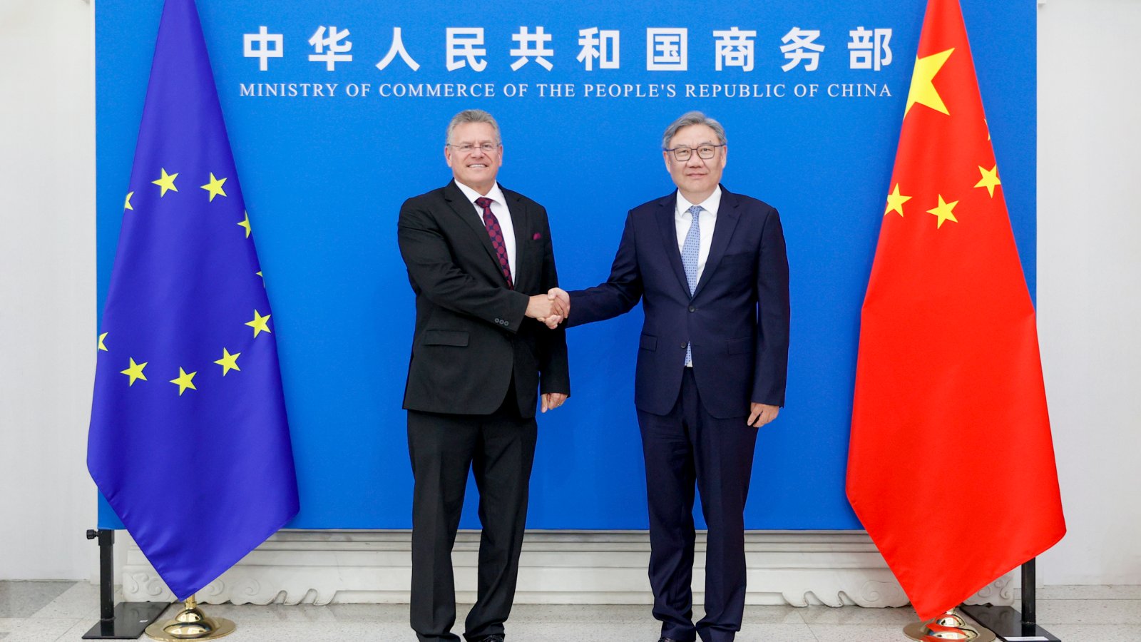 EU Trade Commissioner Maros Sefcovic, left, shakes hands with Chinese Commerce Minister Wang Wentao in Beijing, flanked by EU and Chinese flags.