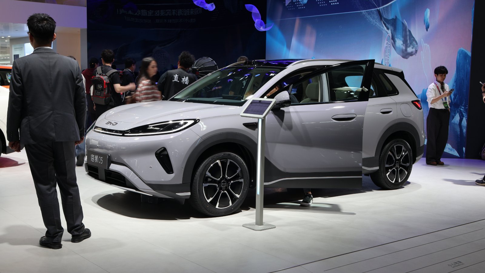 A silver BYD Sealion 05 SUV with its front door open on an auto show floor, as attendees walk past and a man in a gray suit stands nearby.