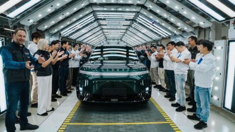 Staff applaud beside a dark green Xpeng G9L in an illuminated factory inspection area.