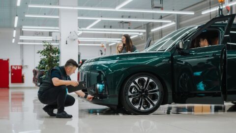 A worker crouches to inspect the front bumper of a dark green Xpeng G9L with its driver’s door open.