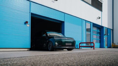 A dark green Xpeng G9L emerges from a factory building with blue doors.