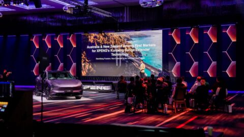 An Xpeng L03 SUV beside a large presentation screen at an event with seated attendees.