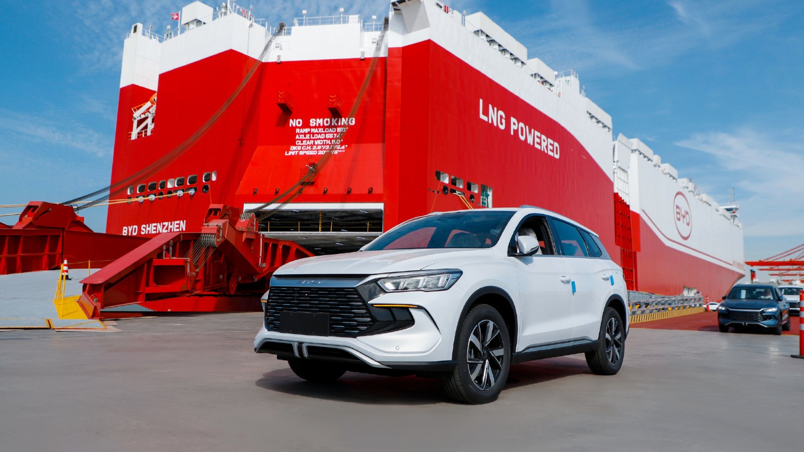 A white BYD SUV beside the red-and-white BYD Shenzhen car carrier at a port.