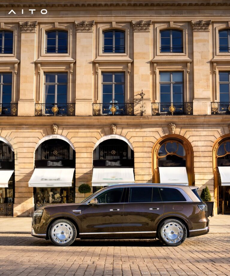 Side profile of a brown Aito 9 SUV in front of a stone building with arched storefronts.