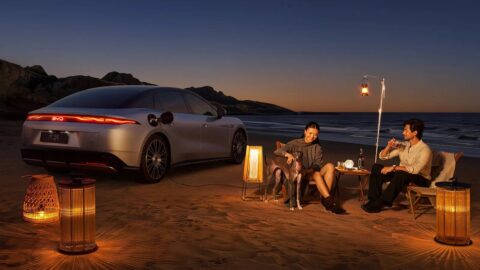 Rear three-quarter view of a BYD Da Han EV beside a beach campsite at dusk, with 2 people seated nearby.