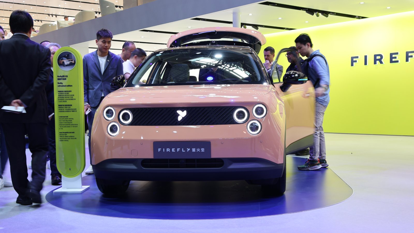 Front view of a peach-colored Firefly EV with illuminated circular headlights, surrounded by visitors at the 2026 Beijing auto show.