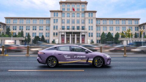 Side view of a purple GAC Aion Ray 7 driving past a large building.