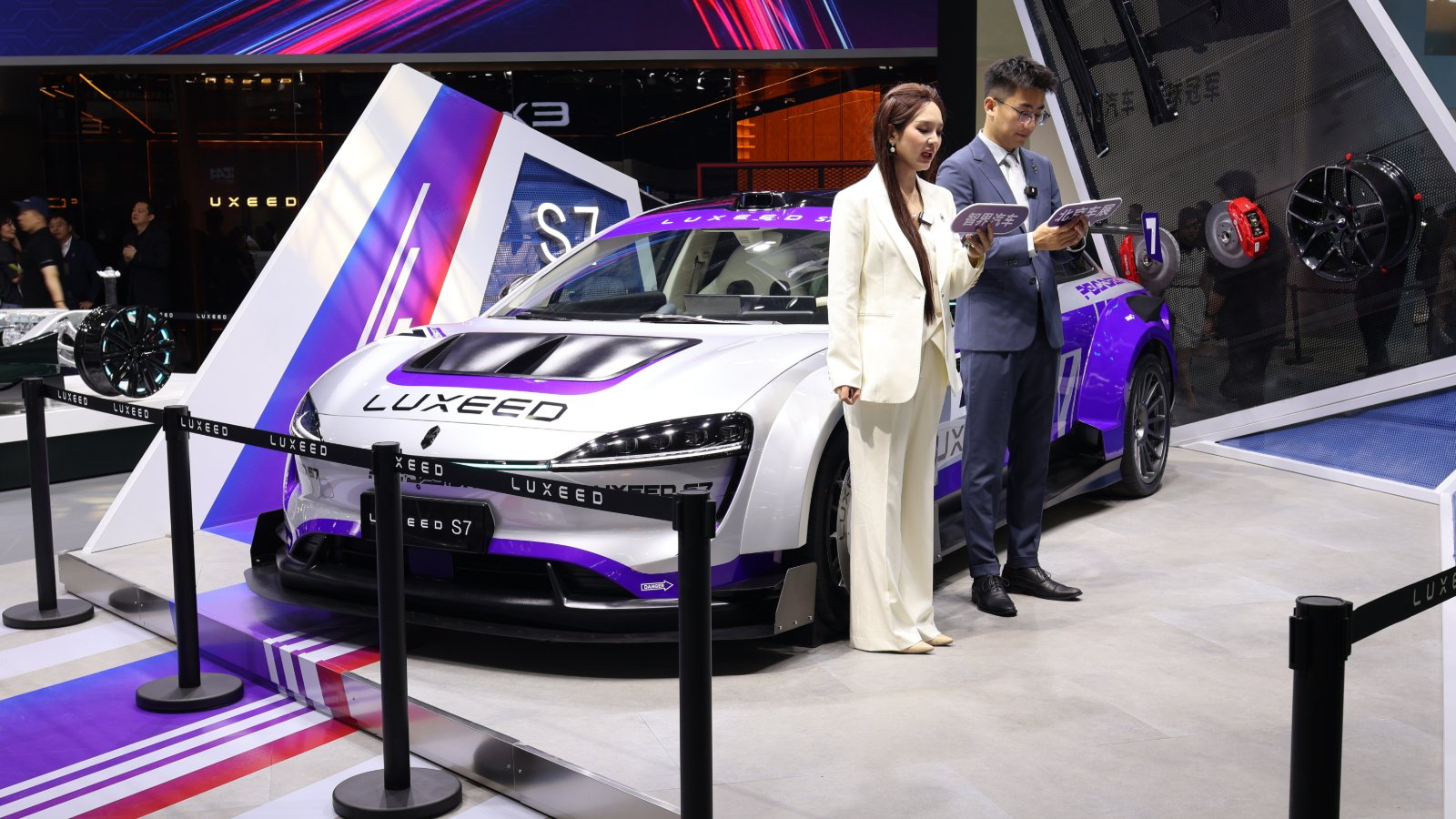 A white and purple Luxeed S7 sedan with racing-style livery on display behind barrier ropes at the Luxeed booth at the Beijing auto show, with a female presenter in a white suit and a male presenter in a blue suit standing next to the car holding tablets.