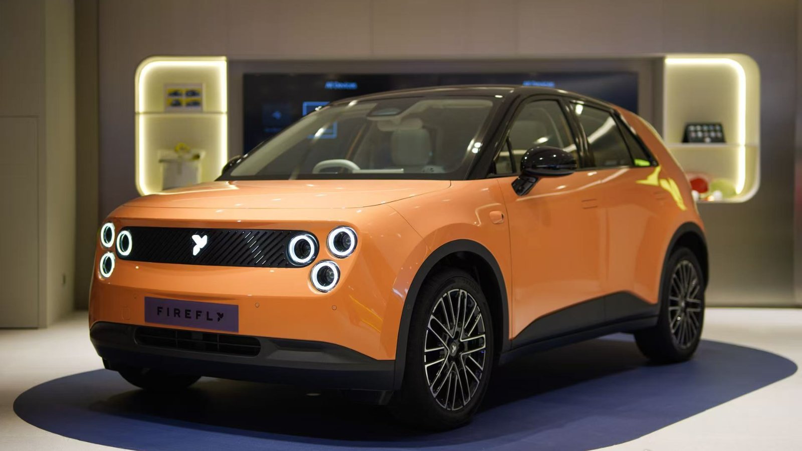 Orange Firefly EV with a black roof and circular headlights on display in a showroom.