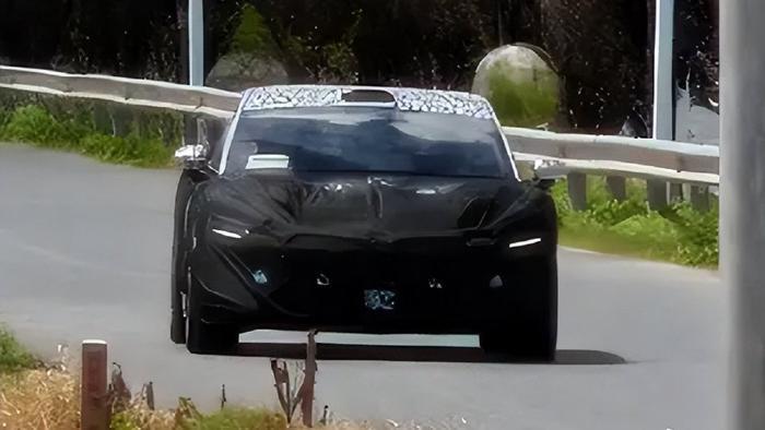 Front view of a camouflaged sedan on a road, with slim illuminated lights and black coverings concealing its bodywork.