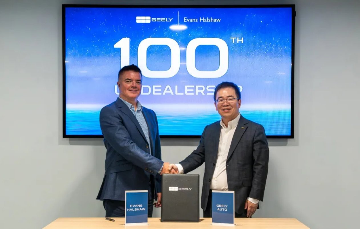 Geely and Evans Halshaw representatives shake hands in front of a display marking Geely's hundredth UK dealership.