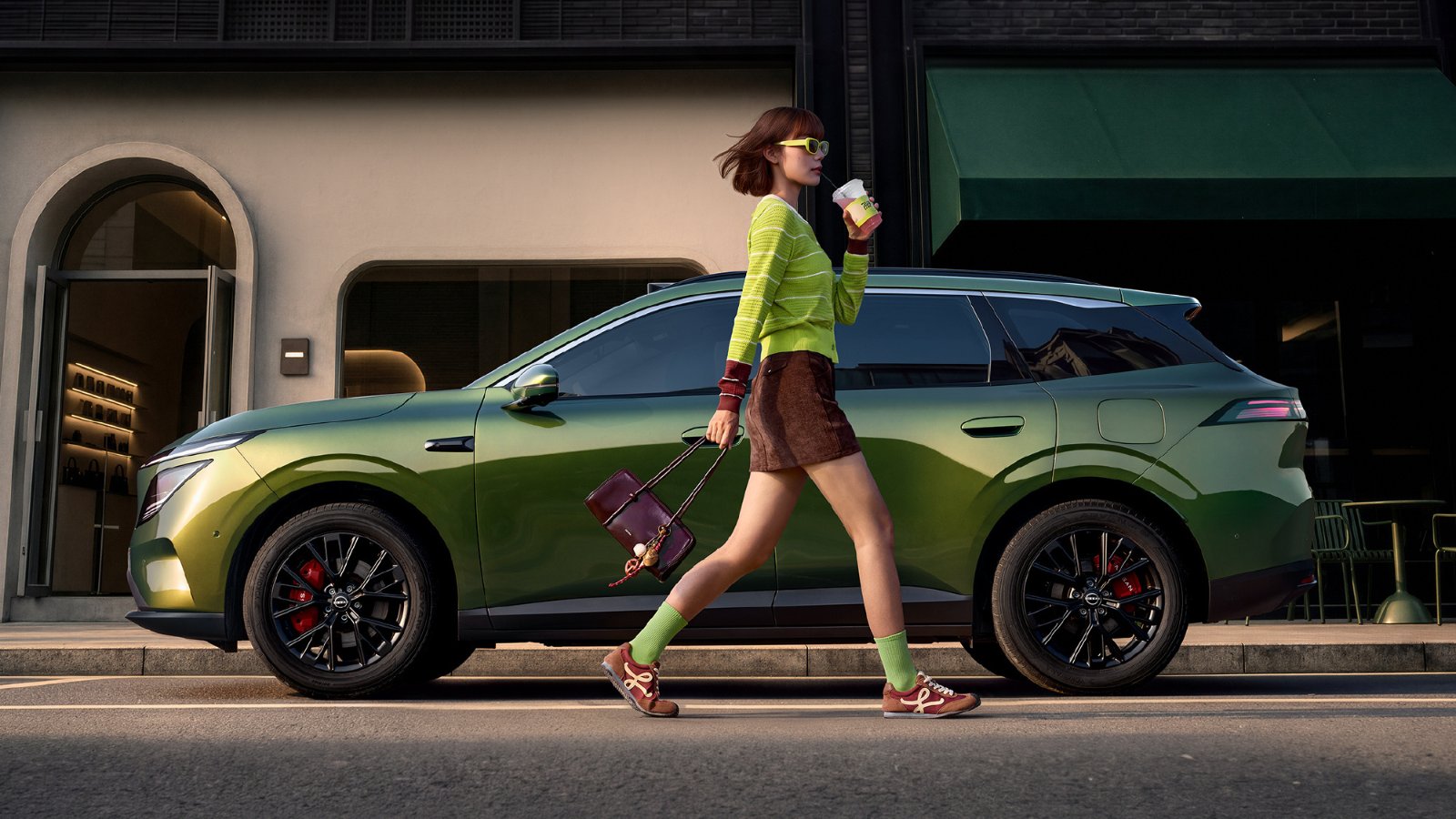 Side profile of a green Dongfeng Nissan NX7 SUV parked along a city street, with a woman in a yellow-green sweater walking past it holding a coffee cup.