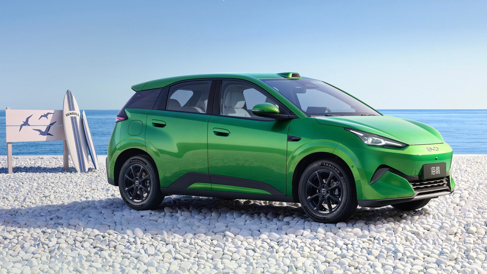 Green BYD Seagull electric hatchback parked on a pebble beach, seen from the front three-quarter angle, with surfboards and a sign reading Seagull on the left and the sea in the background